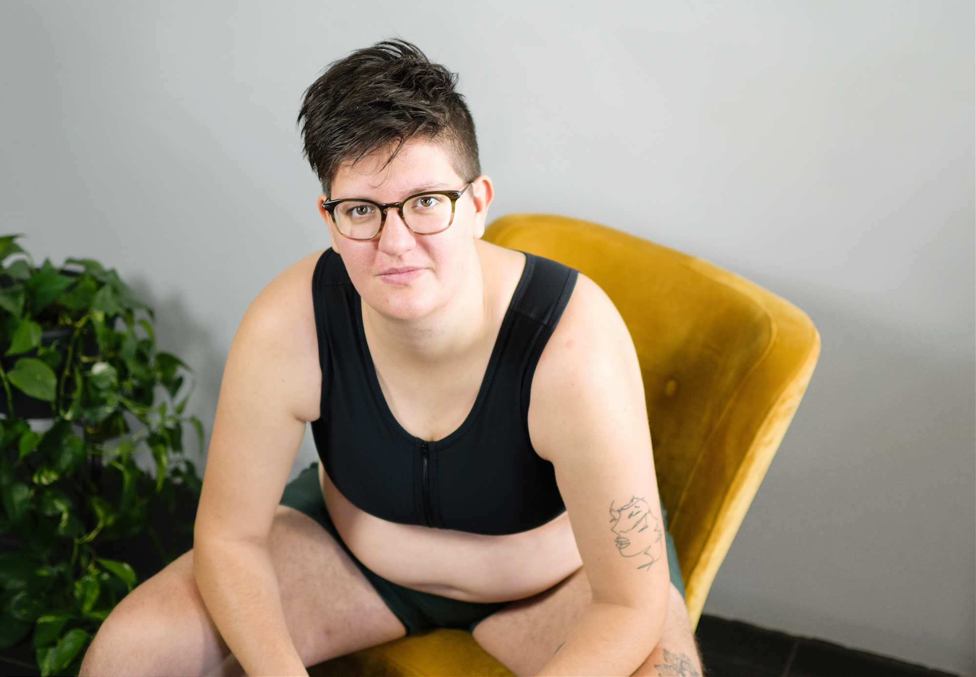 Tilke Wouters sitting on a yellow chair and wearing a short black binder and underwear.
