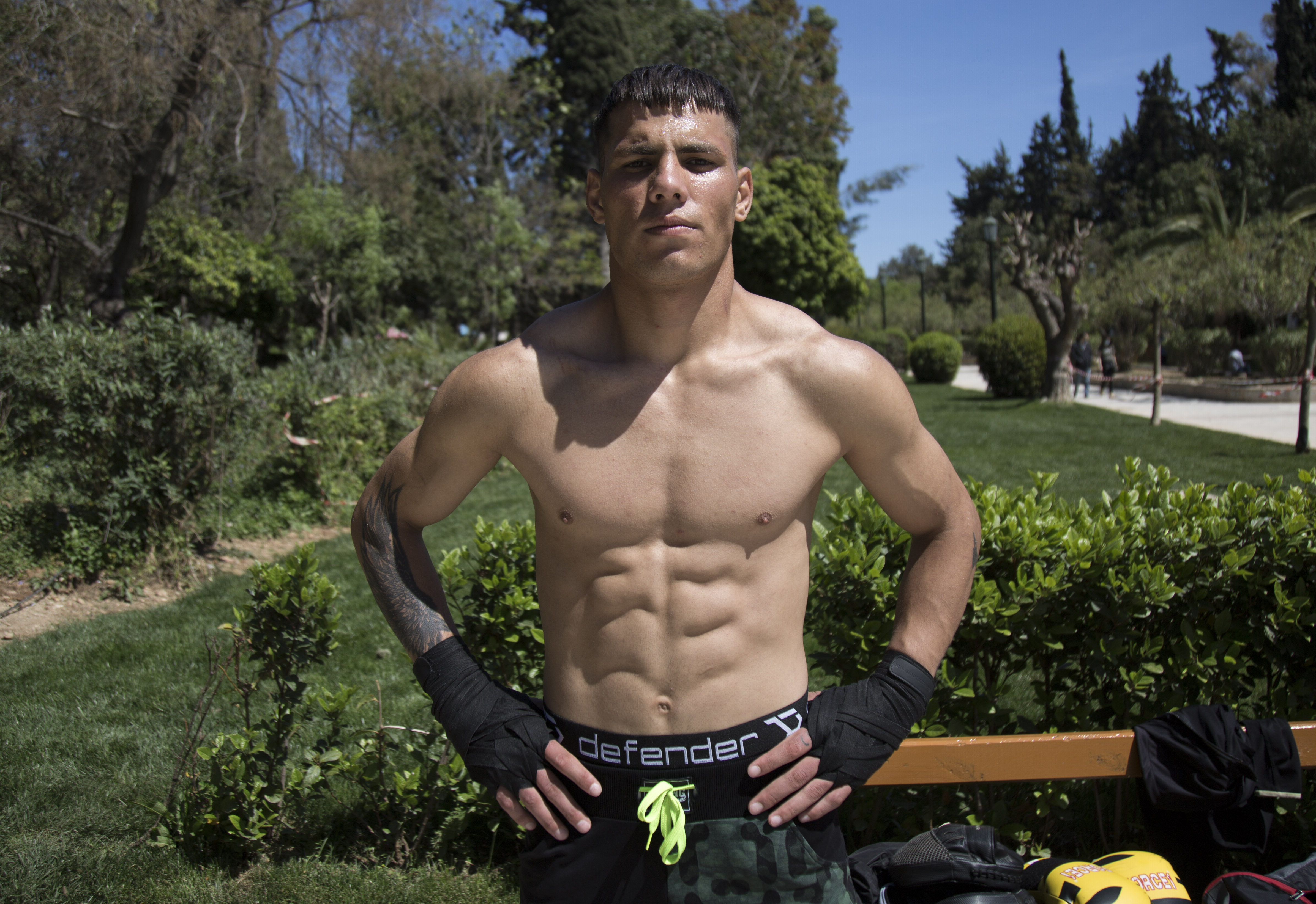 IQBAL ALIKHIL - Muscular teen standing shirtless in a power pose with his hands on his hips, facing the camera. Background: park.