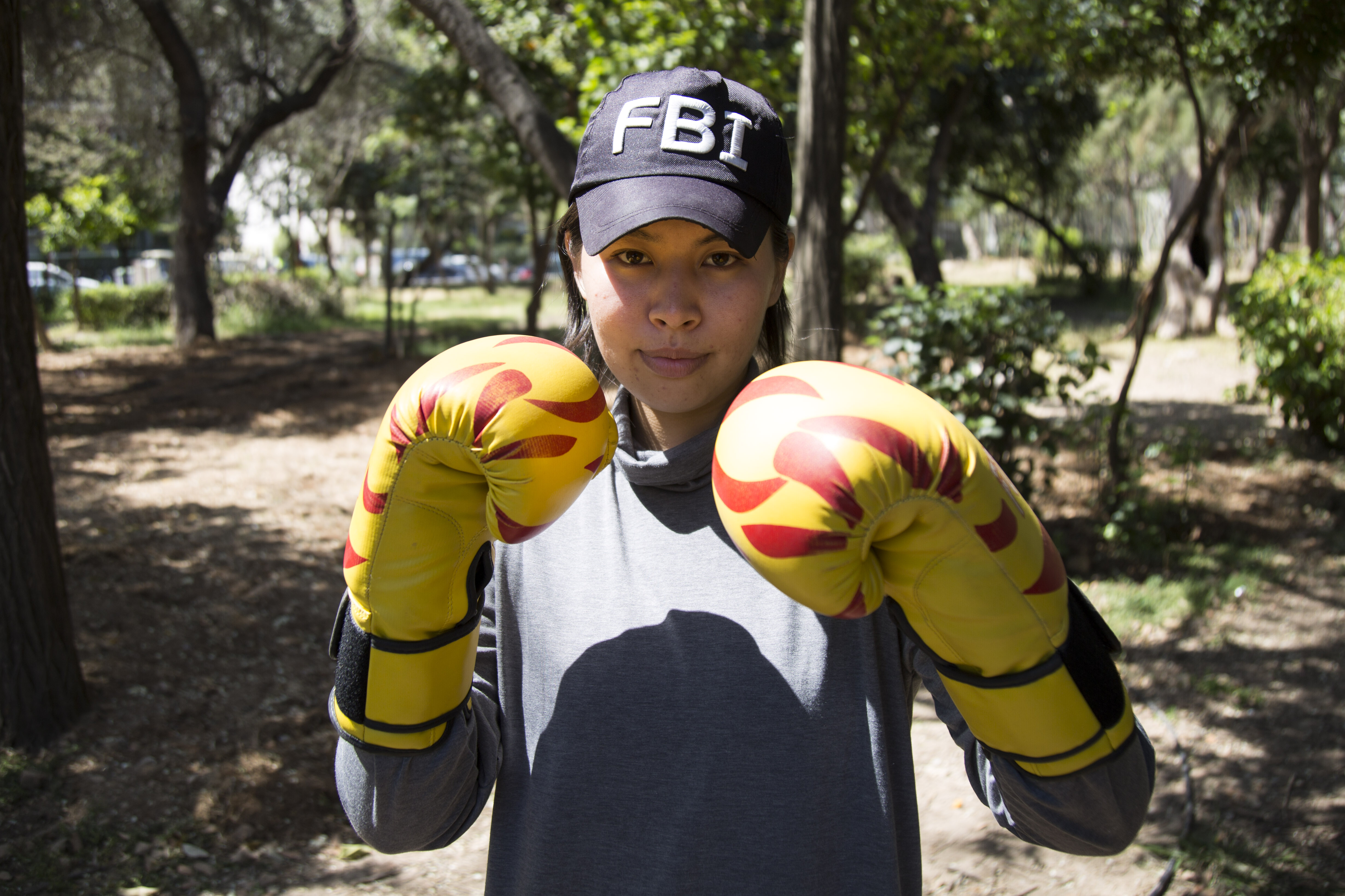 ZAHRA KHWARE - Teen wearing an FBI cap, a gray shirt and yellow and red gloves, holding her fists up to her face and staring at camera.