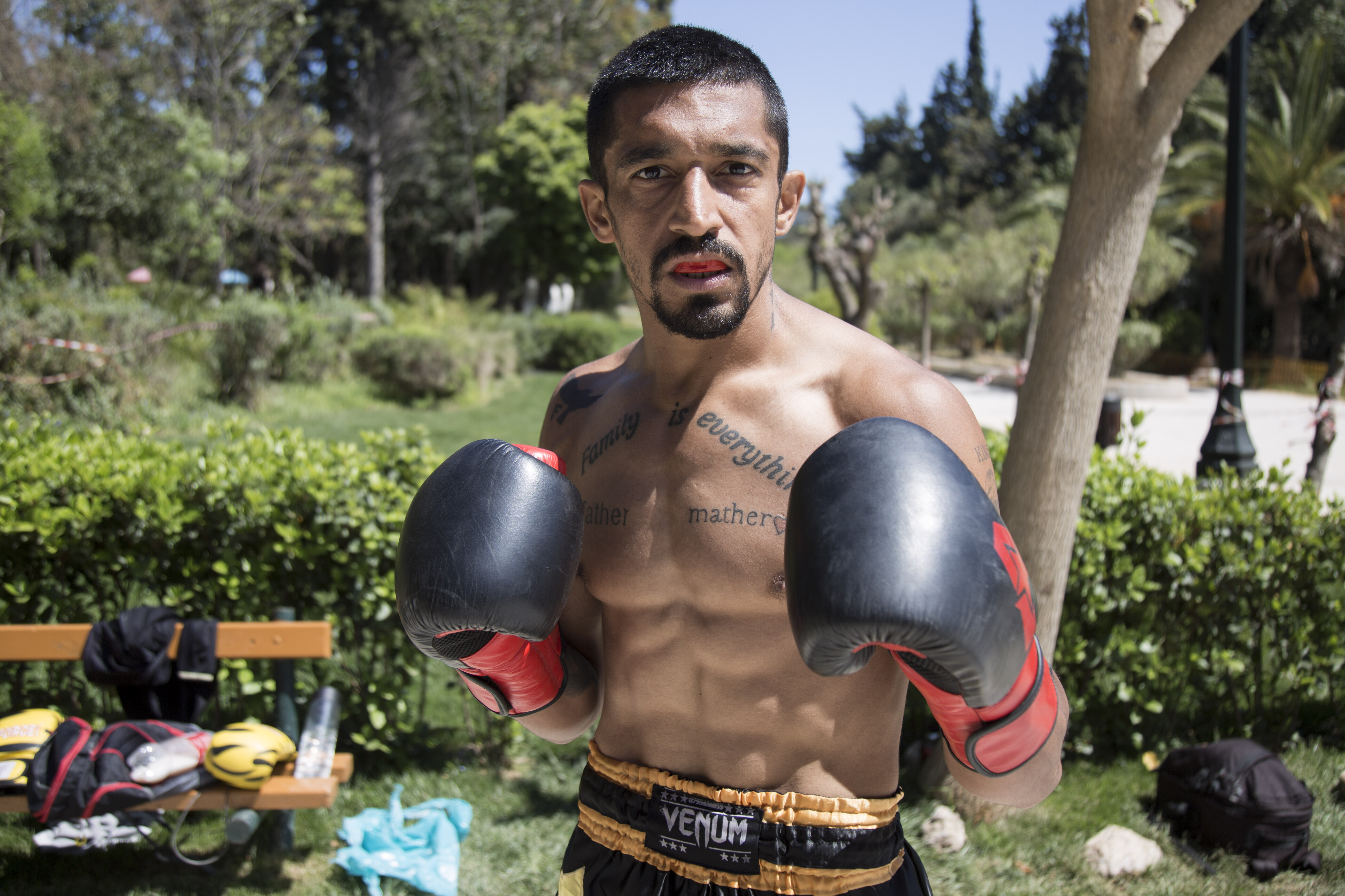 Hamid Norouzi – muscular shirtless man with tattoos wearing a red spit guard, red and black gloves and looking straight at camera.