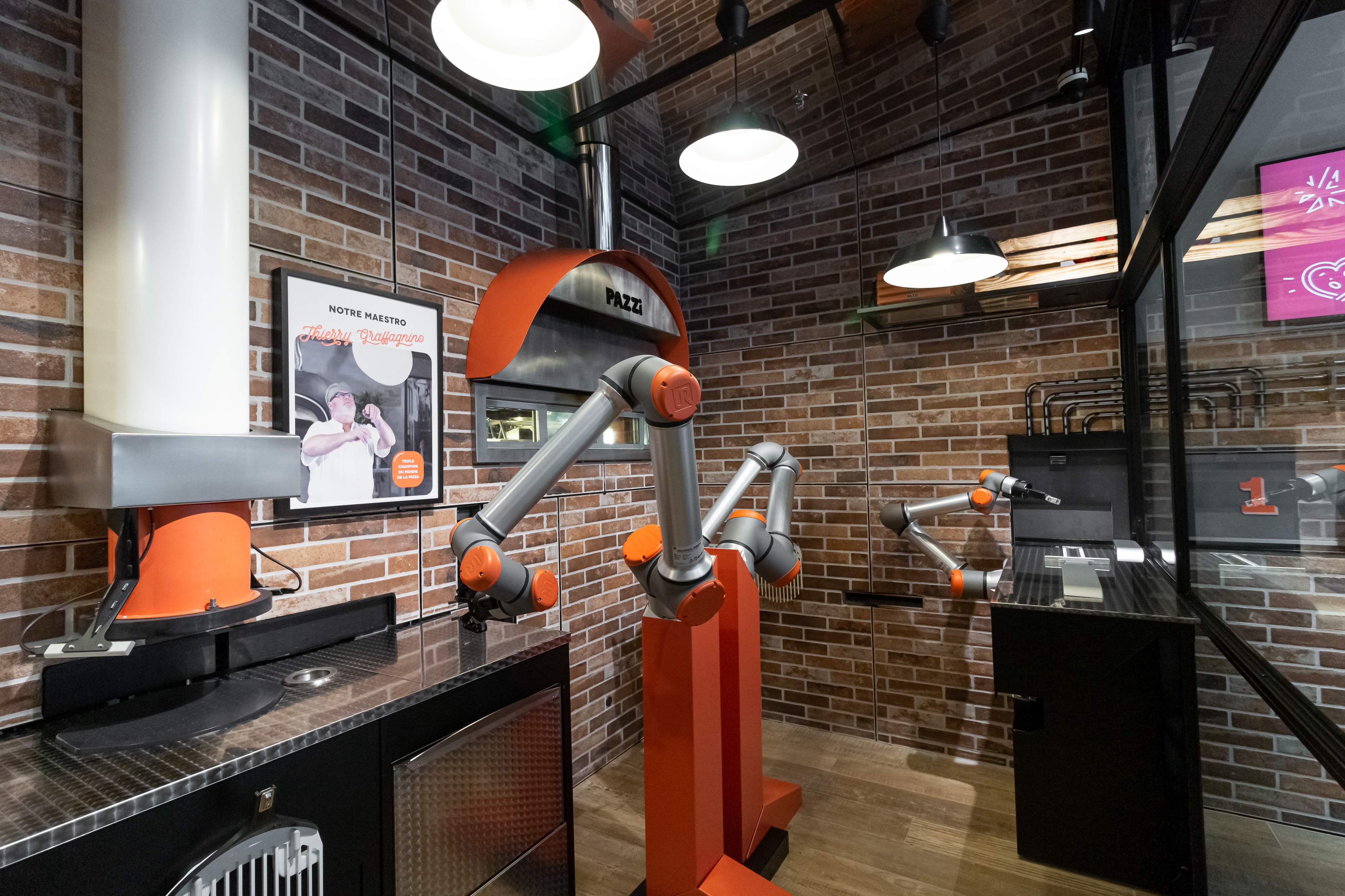 Pazzi pizza robot, Paris – Pazzi pizza robot, Paris – two red and silver mechanical arm robots inside a brick wall kitchen with a large oven.