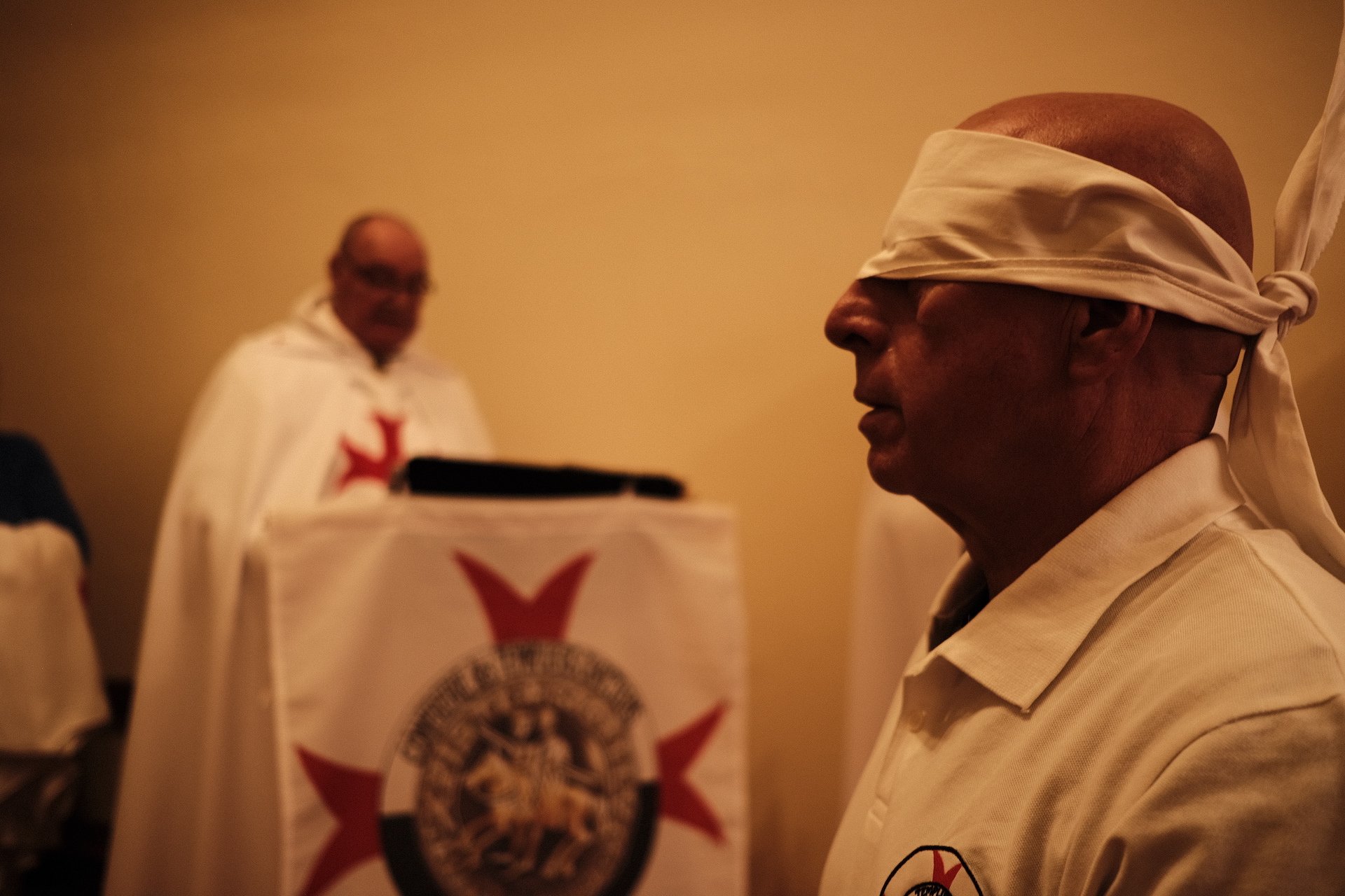 Ein Mann mittleren Alters mit Glatze und weißer Augenbinde, im Hintergrund ein Mann hinter einer Kanzel von der ein Tuch mit Templerkreuz hängt