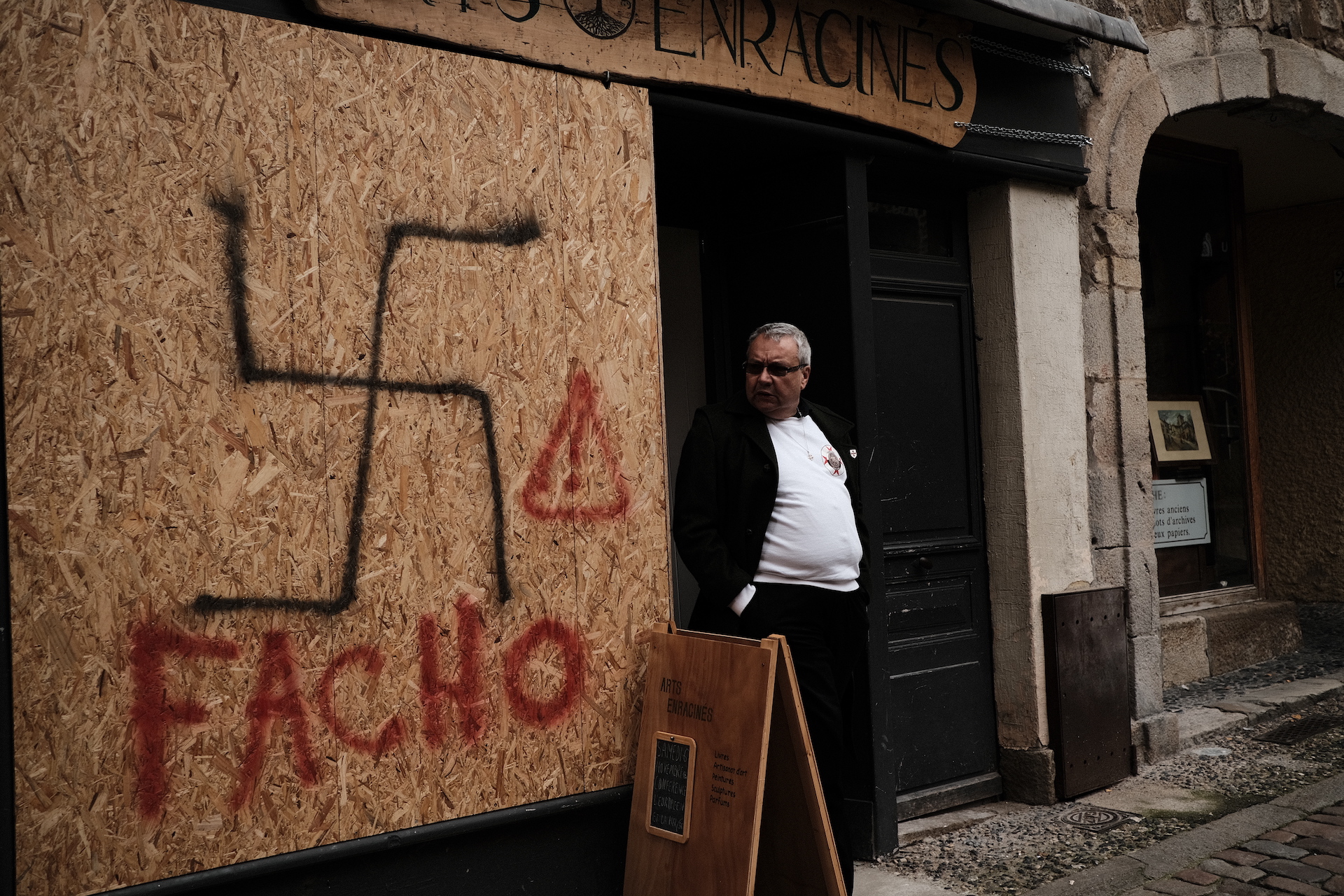 Ein Mann mit grauen Haaren steht neben einer Sperrholzwand, auf der ein Hakenkreuz und das Wort Fascho gesprüht sind