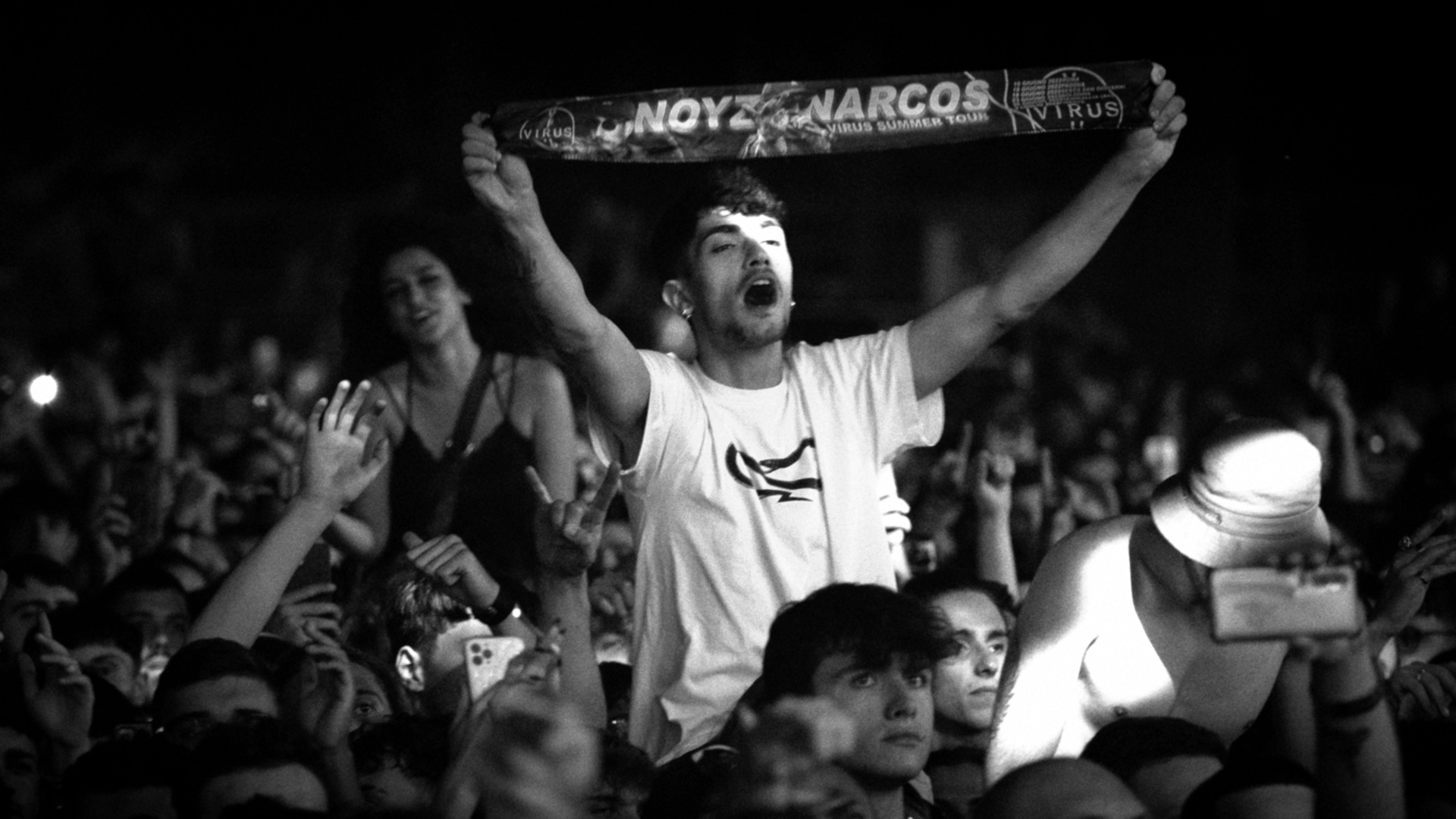 La folla a un concerto di Noyz Narcos. Un ragazzo spicca nel centro con in mano una fascia del tour.