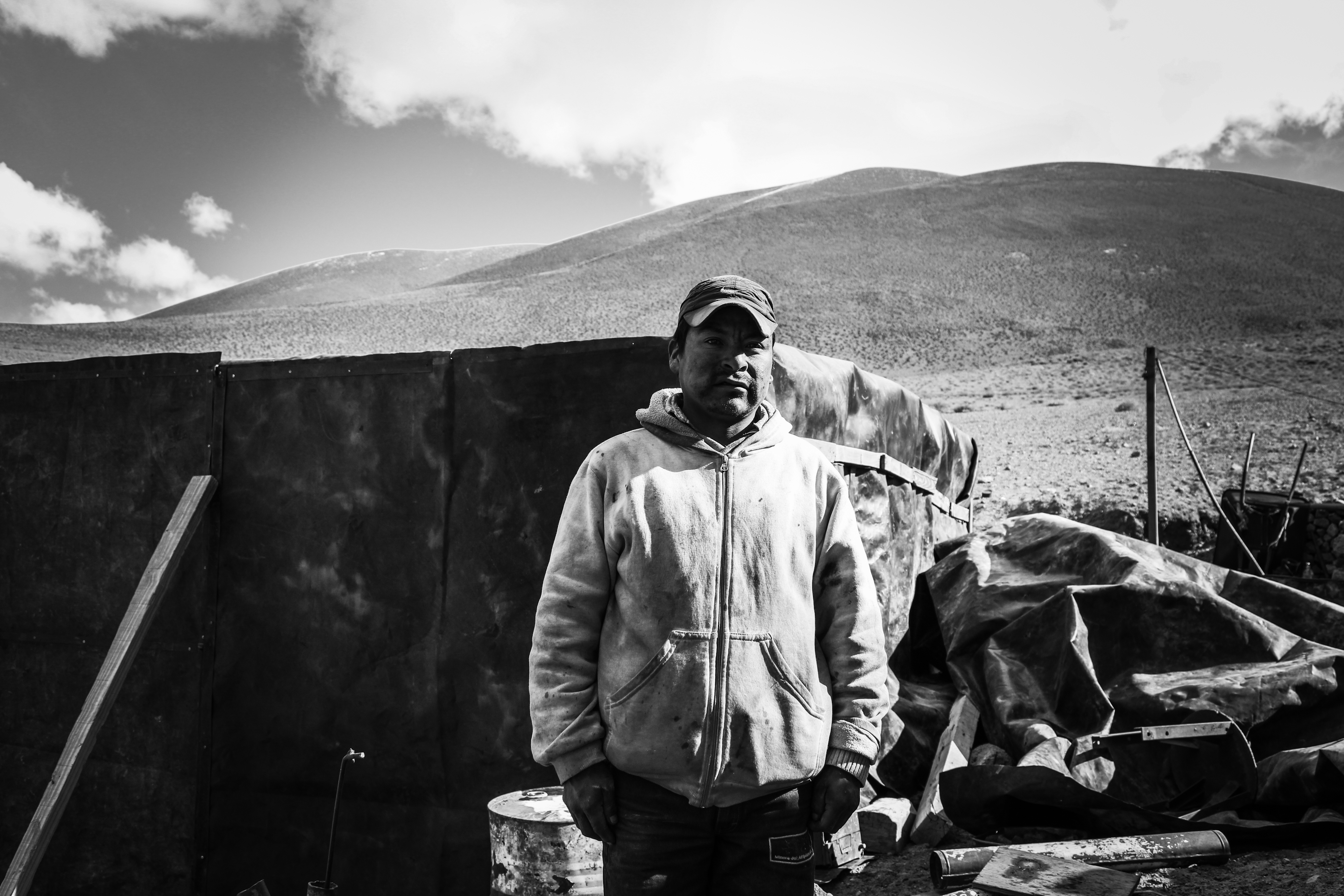 Black and white image of a man in a baseball cap stood in front of a dark wall, mountains visible in the distance.